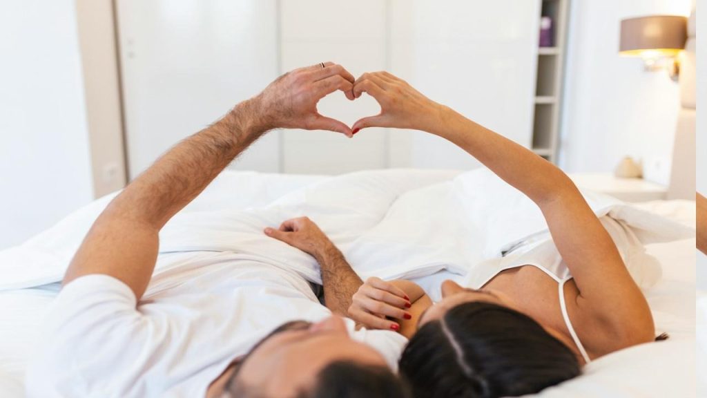 A couple lying on bed forming a heart shape with their hands.