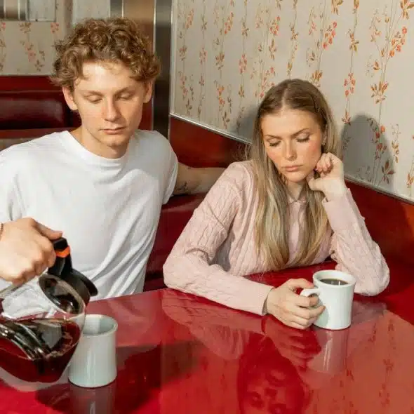 A waiter pours coffee in a mug while a couple is watching silently.