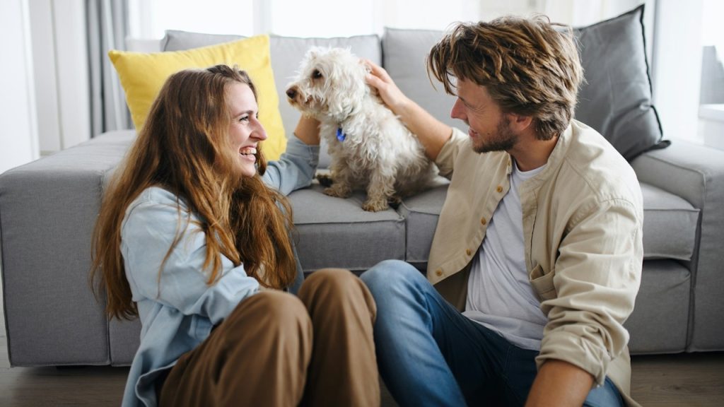 A couple smiling and sitting on the floor while the man strokes a dog who’s sitting on a couch.