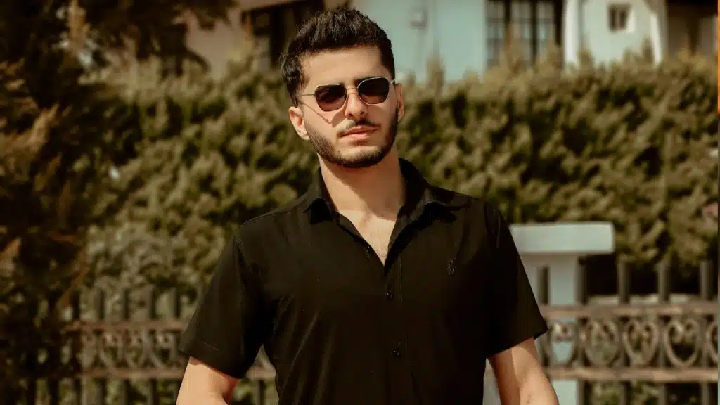 A man wearing a black polo and black sunglasses is walking through a street.