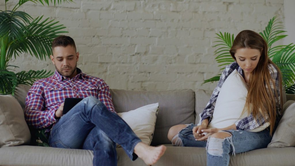 A man using his cell phone while his wife is sitting near him appears upset.