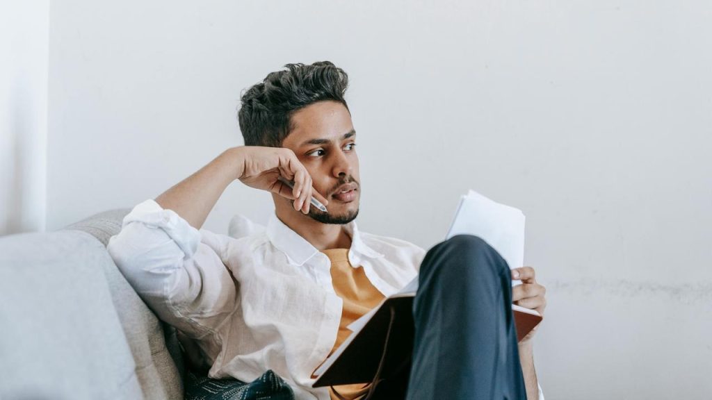 A man sitting on a couch, holding a notebook and looking thoughtfully to the side.
