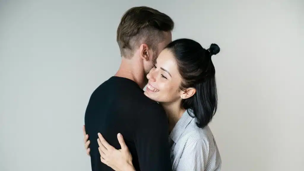 A smiling woman hugs a man from the front with her eyes closed, her face resting against his shoulder.