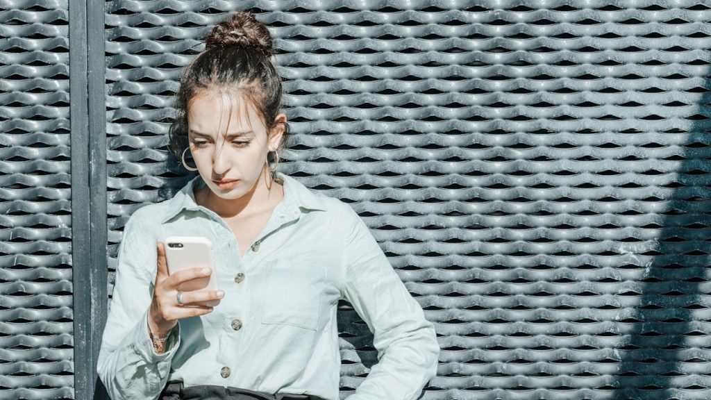A woman is looking at her phone while leaning against a wall.