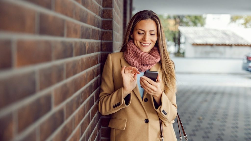 A woman smiles as she looks at her phone while leaning against a wall.