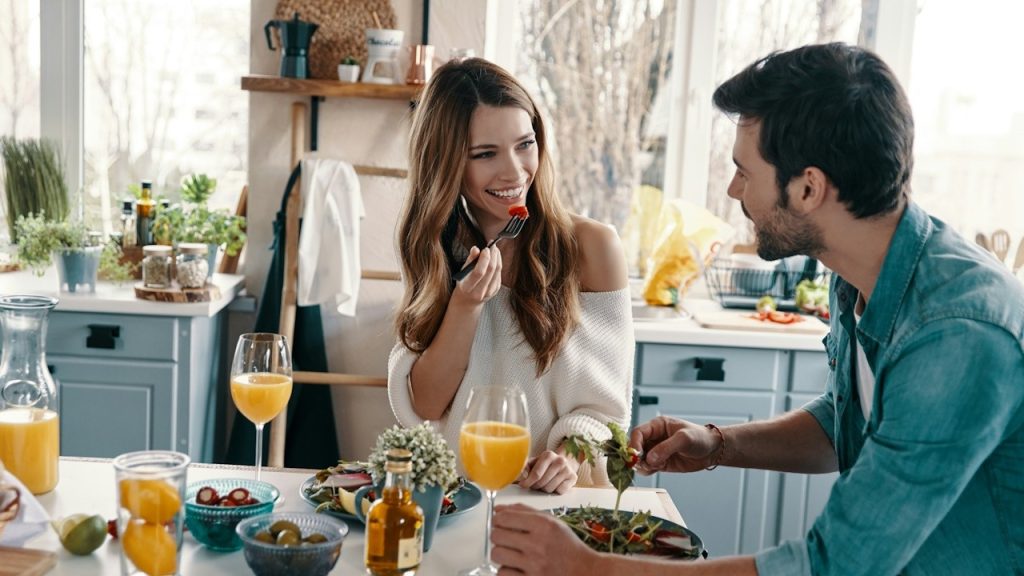 A couple smiling at each other while talking and having dinner together in a kitchen.