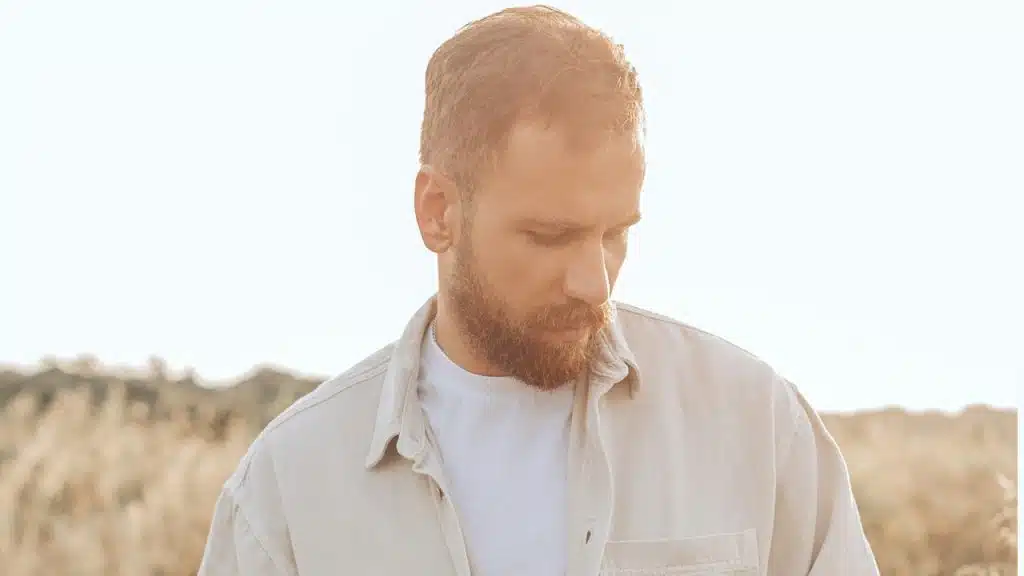 A man wearing a skin-colored off-white shirt is standing in a field.