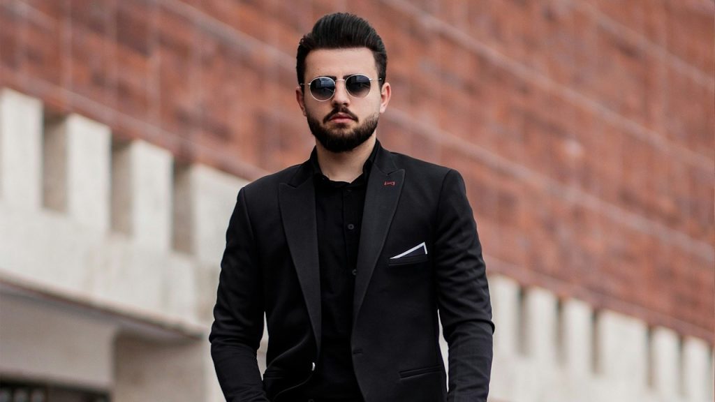 A bearded man wearing a black suit and wearing sunglasses.