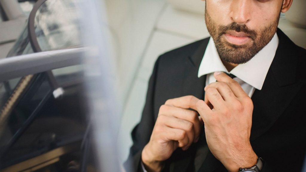 A lightly bearded man adjusting his suit’s tie.