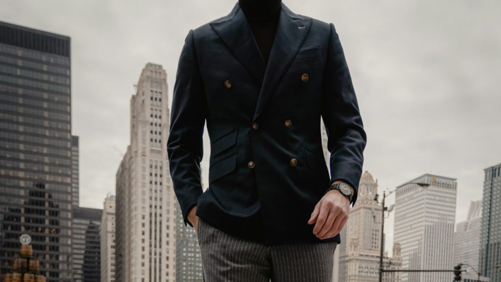 A man in an olive coat standing on the sidewalk during the daytime.