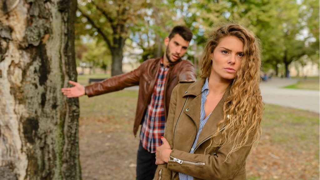 A woman with long wavy hair, wearing a brown jacket, is looking directly at the camera while a man gestures in the background near a tree.