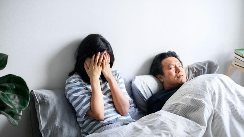 A stressed woman with hands on her face is sitting in bed with a man sleeping beside her.