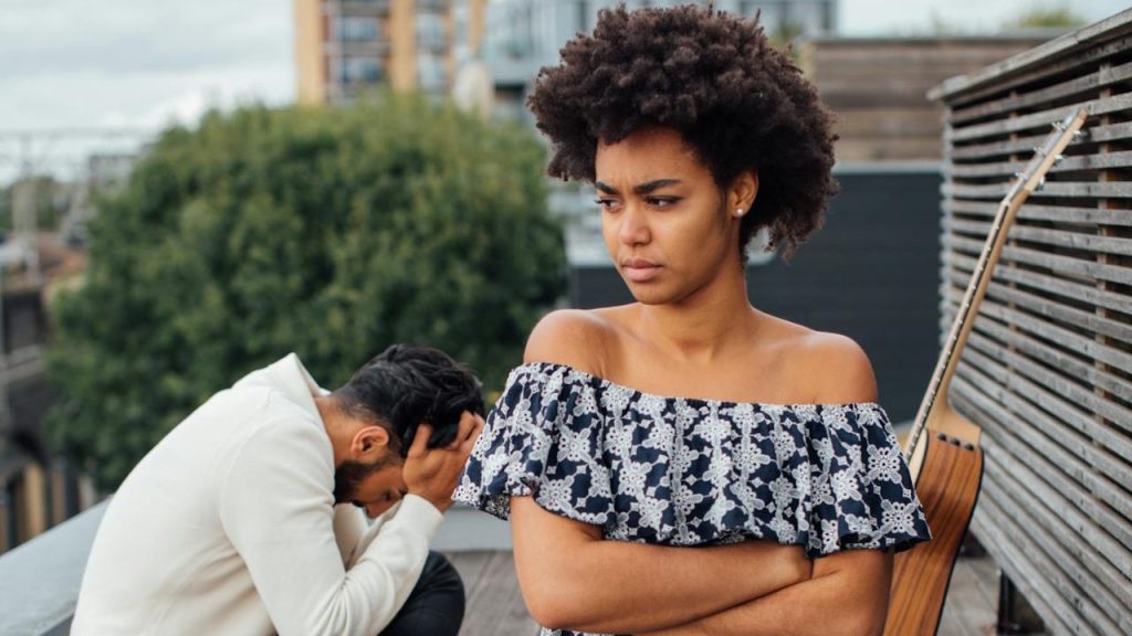 A Black woman with arms crossed, kneeling and looking upset on a rooftop picnic while a man holds his head in the background.
