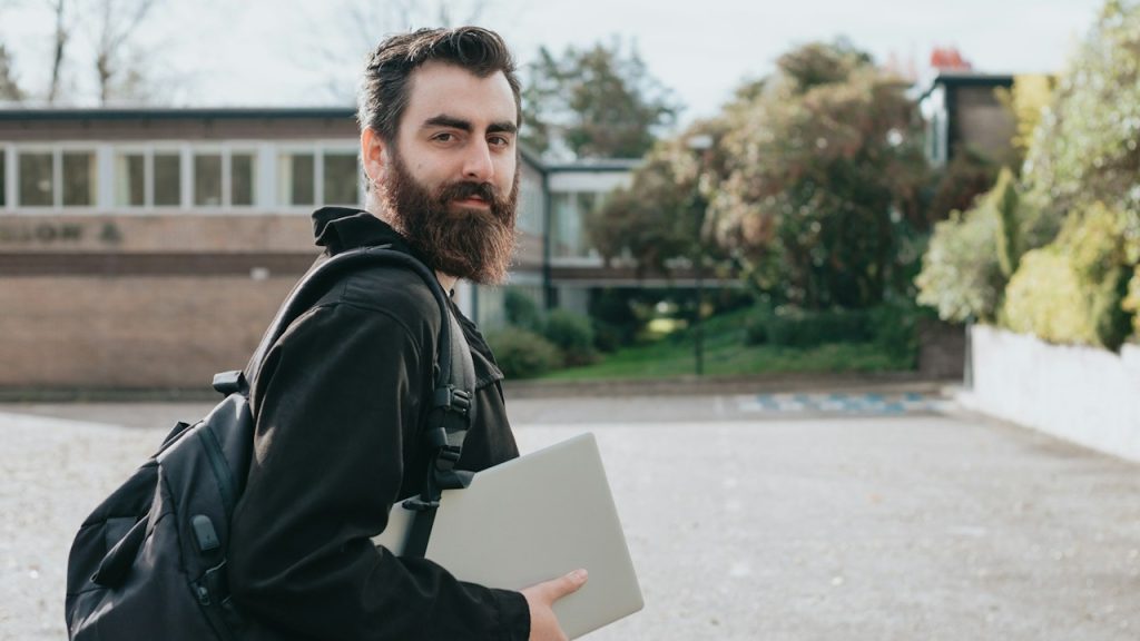 A man with beard is holding a laptop is standing in front of a building.