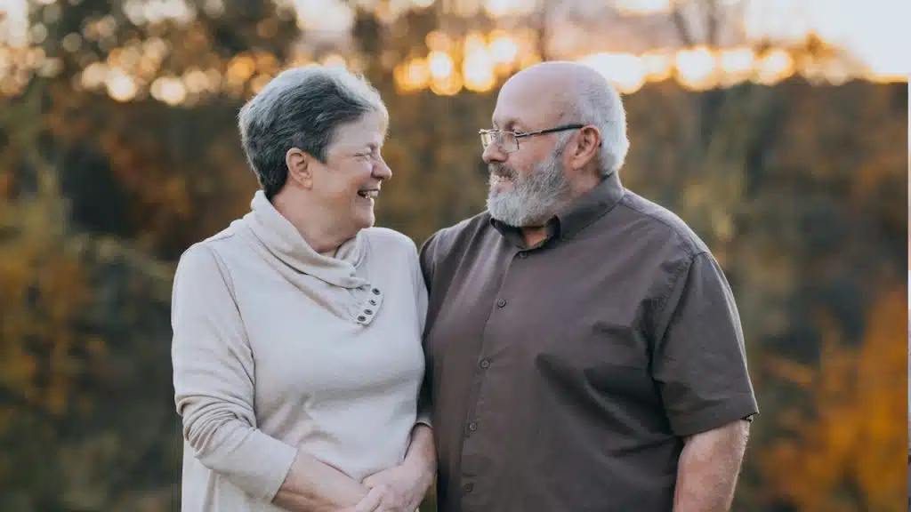 An older couple standing outdoors and smiling at each other. 