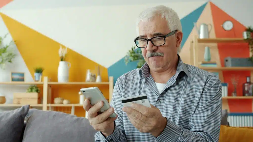 An older man with glasses holding his phone while he looks at his credit card. 