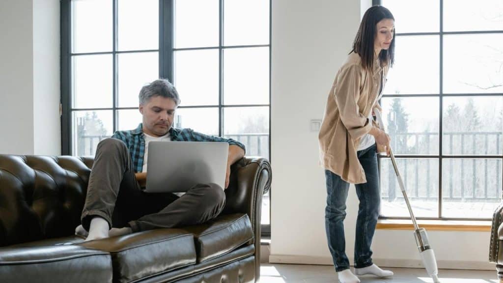 A man uses a laptop on a leather sofa while a woman vacuums the floor nearby.