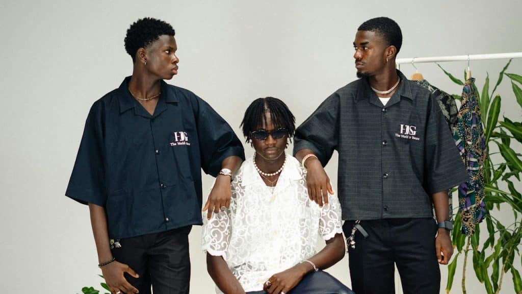 A man in a white shirt is sitting on a stool, and two men in black dresses are standing on his sides.