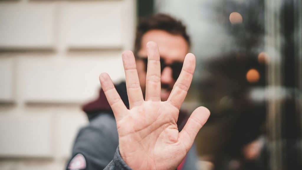 A close-up of a hand held up with five fingers spread, palm facing forward, with a blurred man in the background.