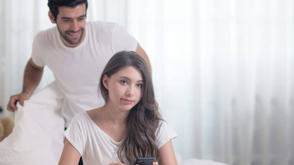 A woman sits on a bed and uses her phone while looking uneasy as a bearded man smiles and approaches her from behind.