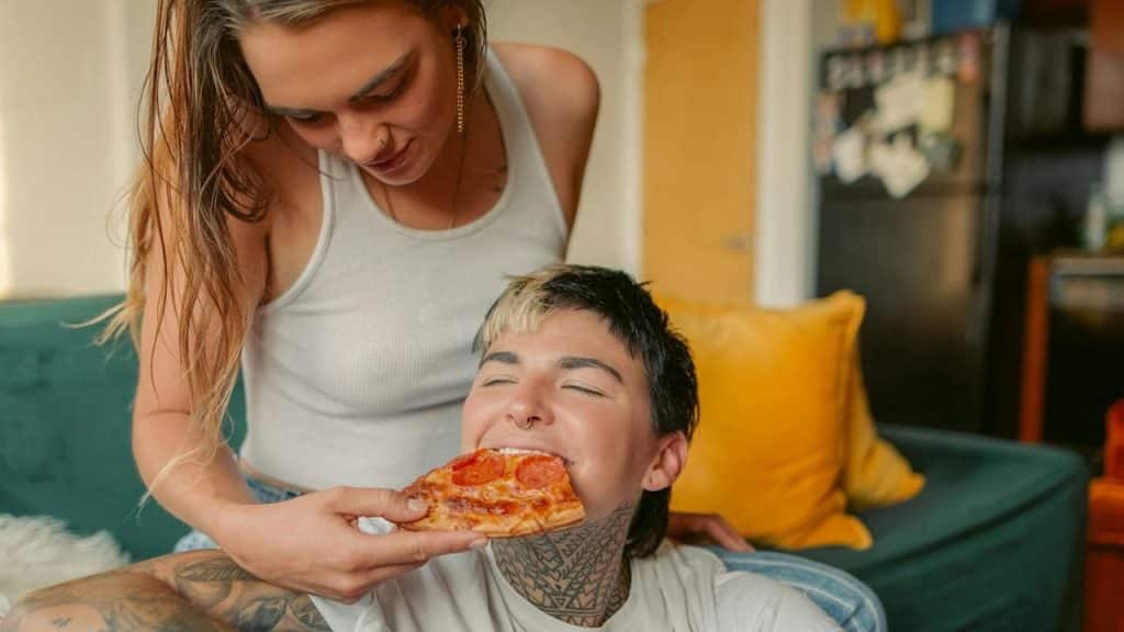 A woman is sitting on a couch feeding a slice of pizza to a man who sits while leaning against her legs.