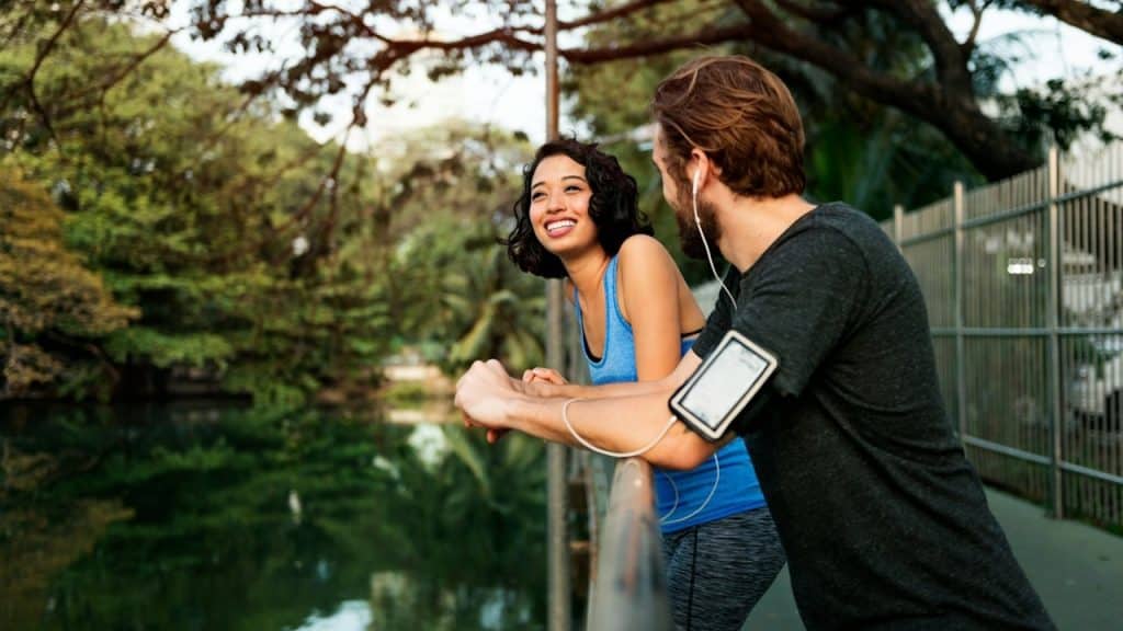 A bearded man smiling and looking at a smiling woman as she leans on a railing over a body of water in the outdoors.