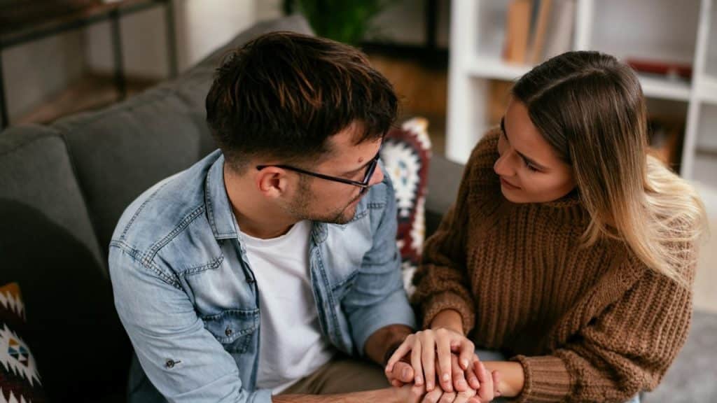 A couple reconciling while holding hands and looking into each other’s eyes as they sit on a couch.