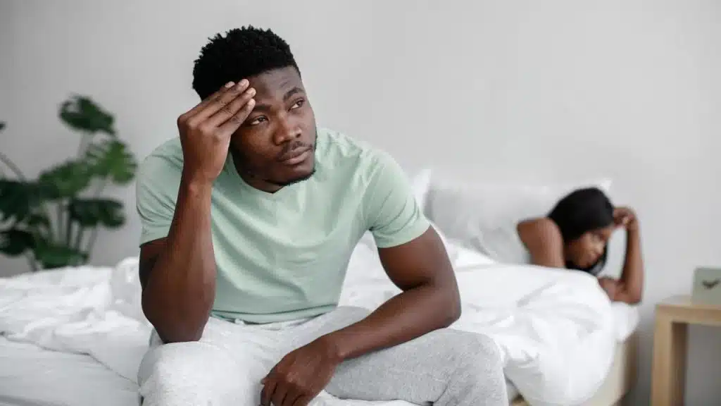 A man sits on a bed looking stressed with his hand on his forehead, while a woman lies facing away from him in the background.