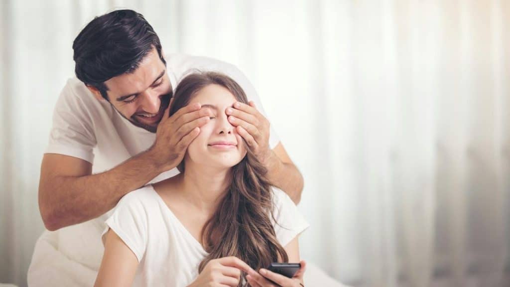 A smiling man covering his wife’s eye.