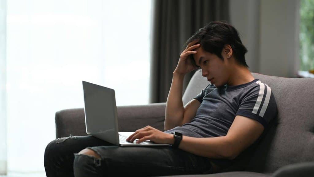 A man sitting on a couch, looking stressed while using a laptop.