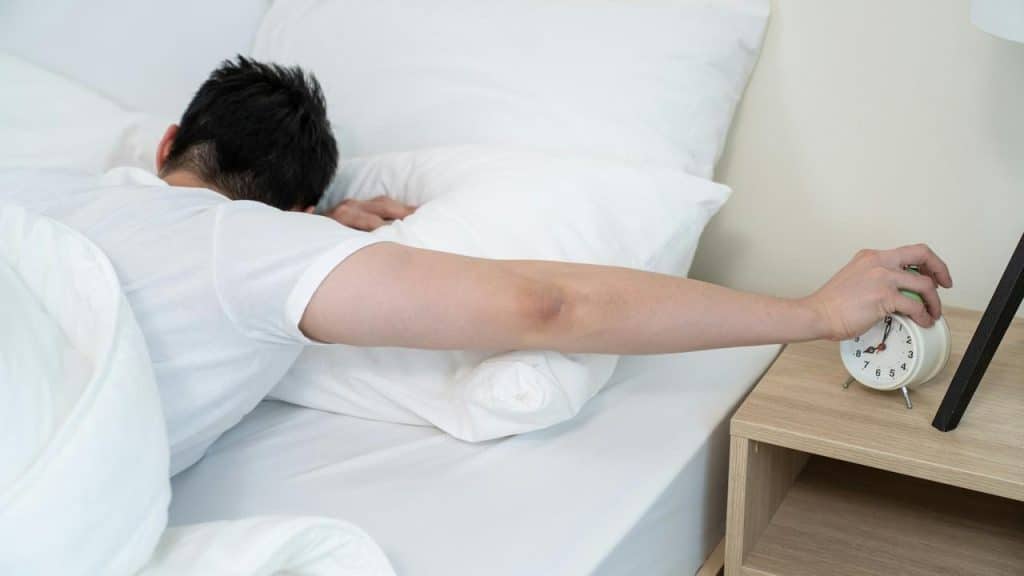 A man lying in bed reaching to turn off an alarm clock.