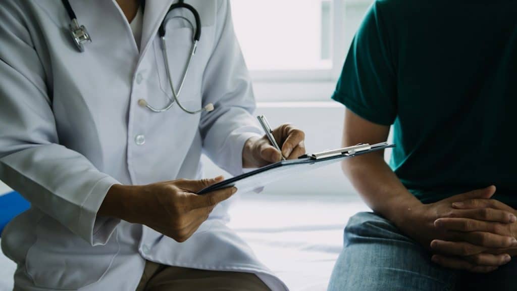 A doctor sitting with a patient and taking notes on a clipboard.