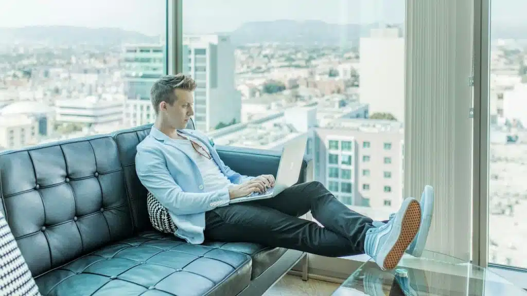 A man in a light blue blazer and sneakers sitting on a black leather sofa, working on a laptop.