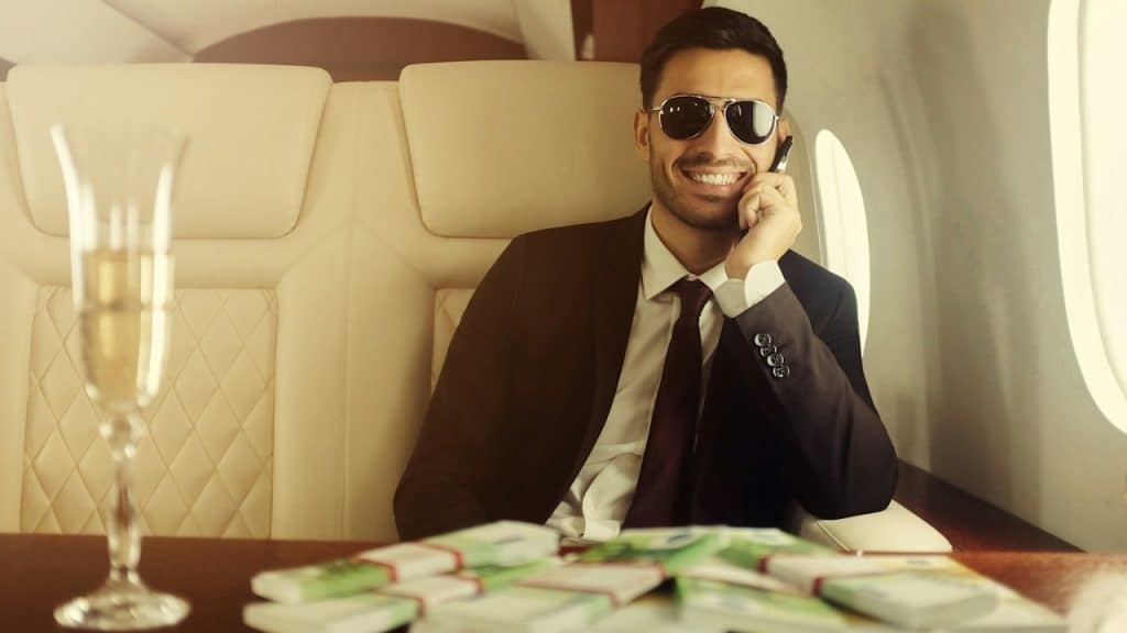 A man in a private jet, with stacks of cash and a glass of champagne on the table in front of him.