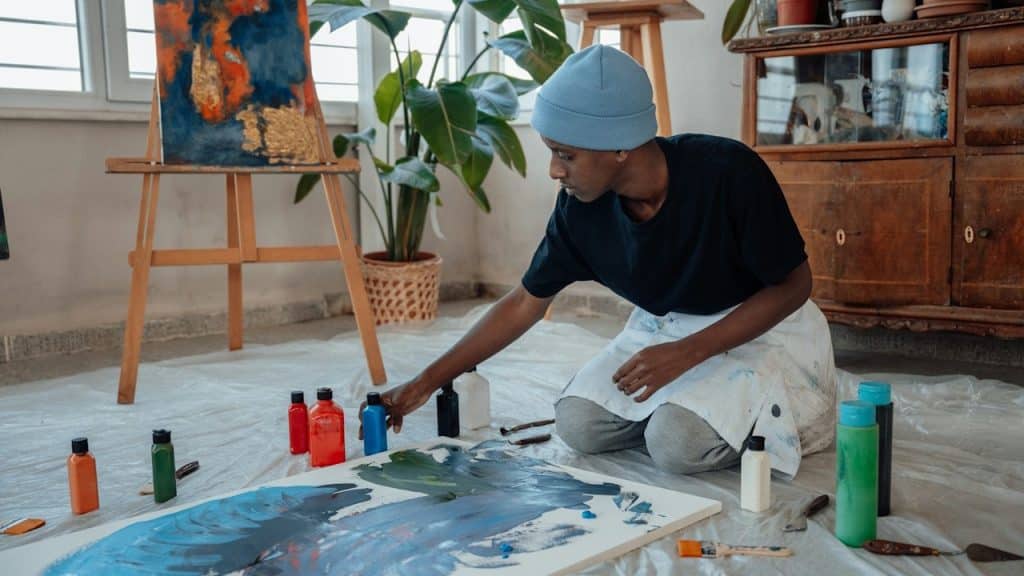 An artist kneeling on a plastic-covered floor, working on a large abstract painting.