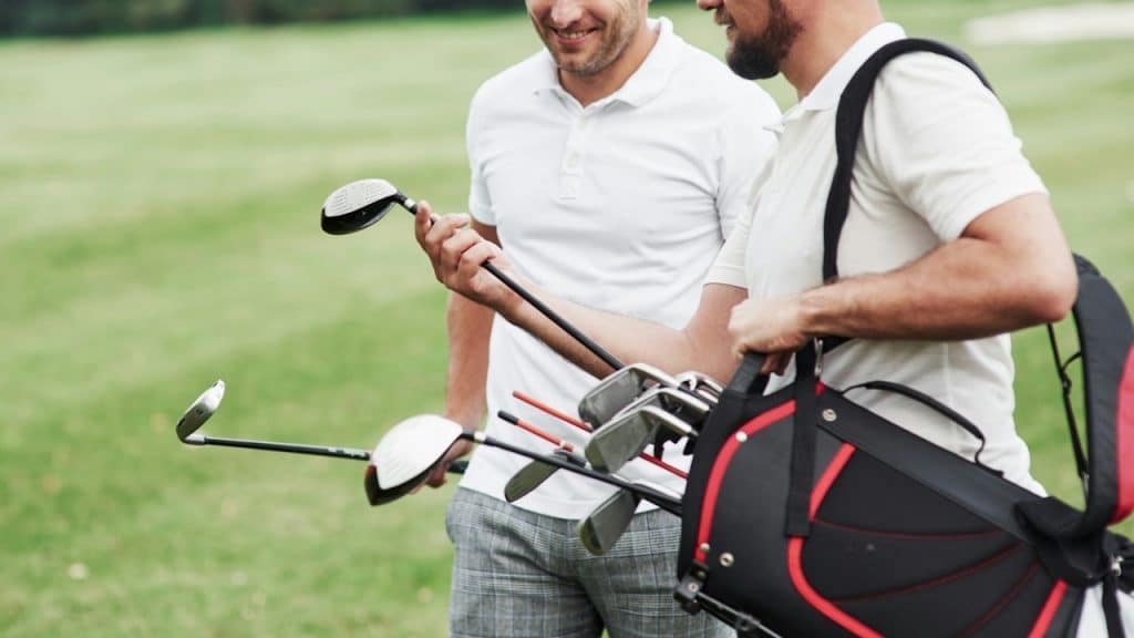 Two men on a green golf course, with one holding a golf club and the other carrying a golf bag.