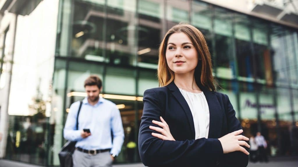 Confident young woman in a dark blazer standing with arms crossed in front of a modern glass office building, with a blurred man in the background.