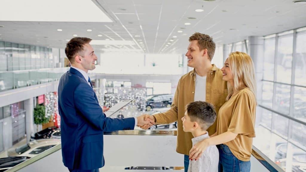 Smiling family of three shaking hands with a car salesman in a bright, modern dealership showroom.