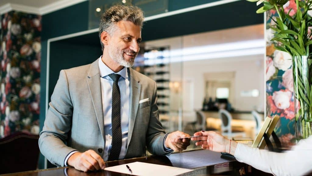 Smiling man in a grey suit handing a credit card to a receptionist.