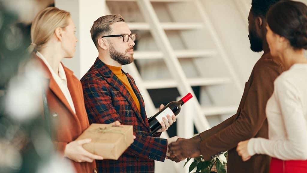A man in a plaid blazer holding a bottle of wine shakings hands with a host while arriving at a dinner party with a guest carrying a wrapped gift.