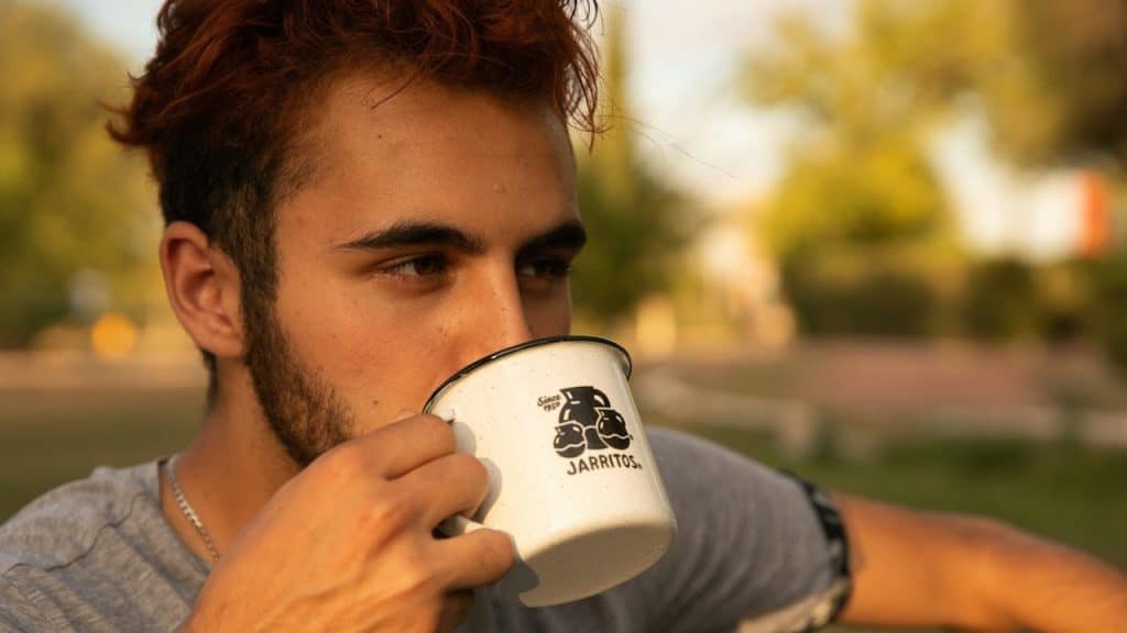 Young man with reddish-brown hair drinking from a white Jarritos enamel mug outdoors.