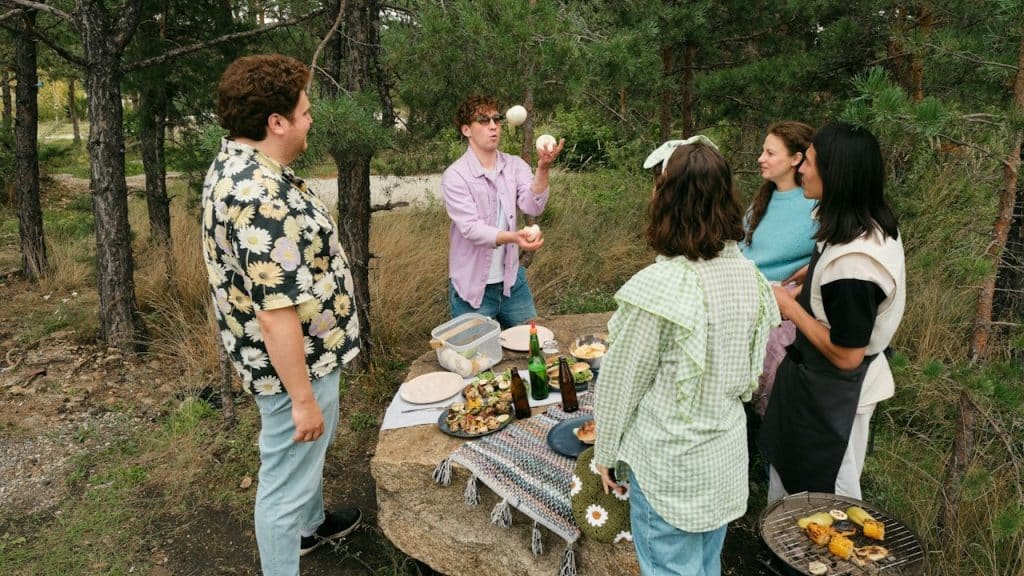"A man juggling onions to entertain his friends during a casual outdoor barbecue in a pine forest."