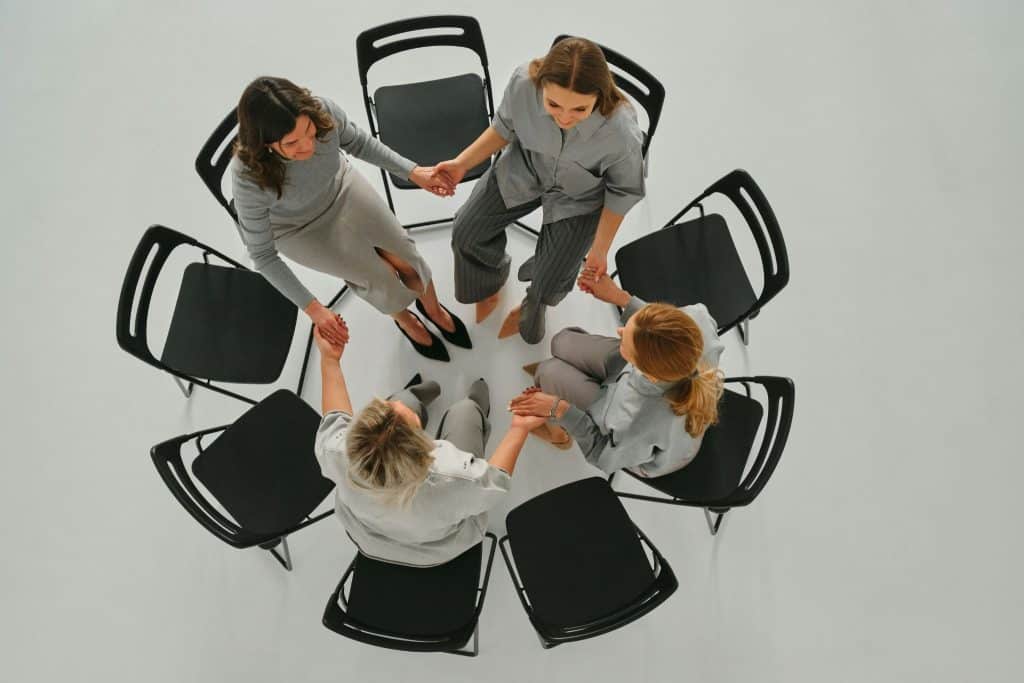Four women sit in a circle on black chairs, holding hands.
