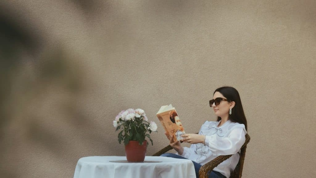 A woman wearing sunglasses reading a book at a table with a flower pot.