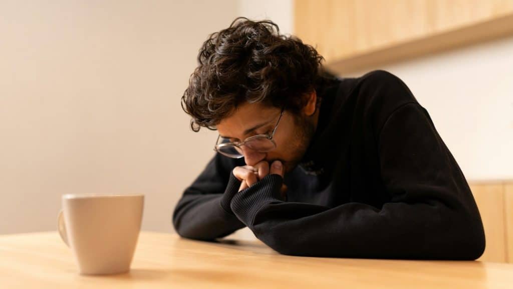 A man with glasses sitting at a table, looking down with a pensive expression, next to a white mug.