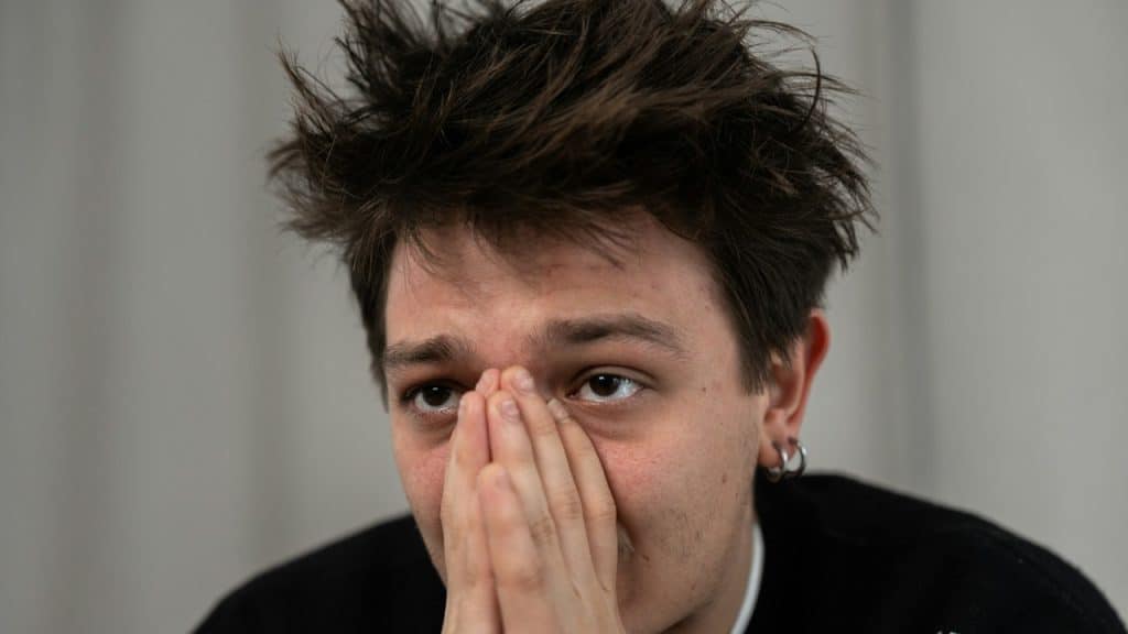 A close-up of a young man with messy hair covering his mouth and nose with his hands, looking worried or anxious.