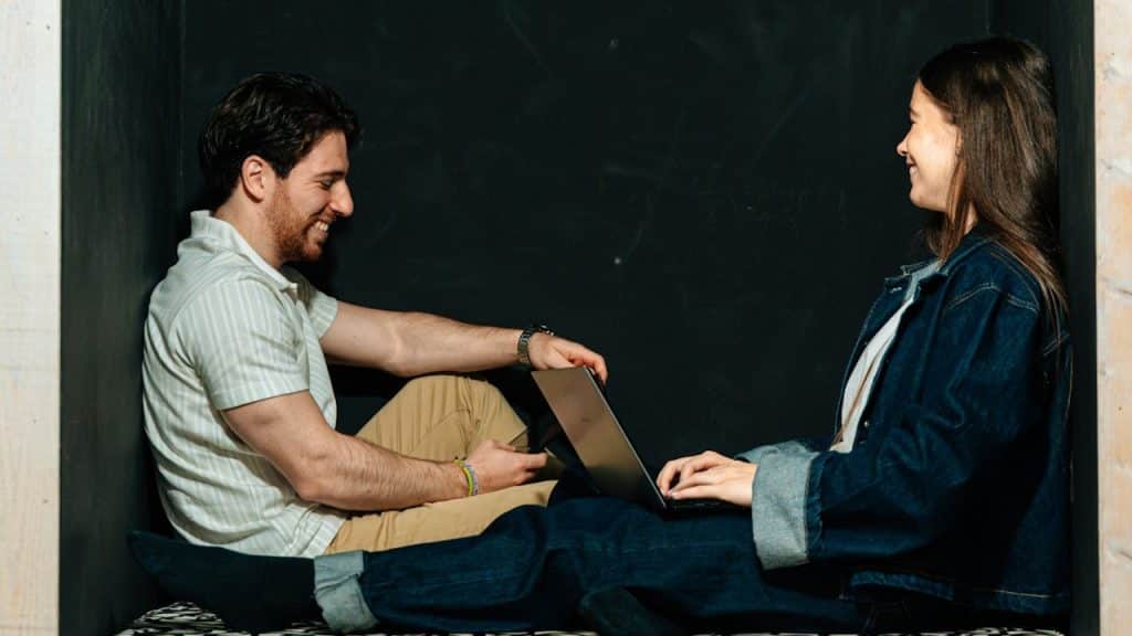 A man and women sitting while women holding a laptop on his knees and both are smiling.