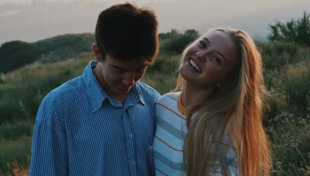 A smiling woman standing close to a man who is looking down as they stand outdoors.