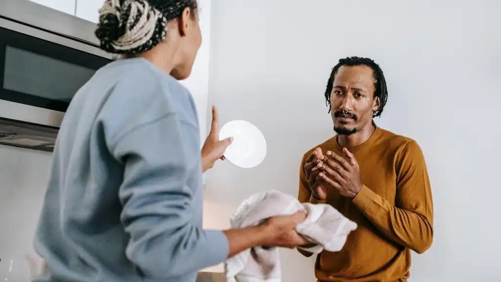 A woman is holding a plate and towel, gesturing while speaking to a man who looks distressed