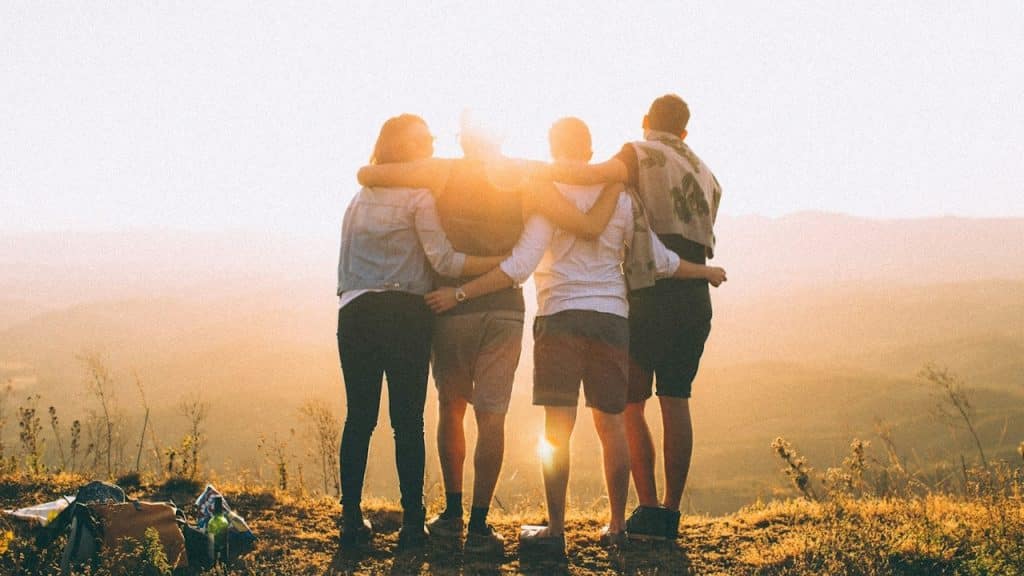 A group of friends standing together with arms on each other's shoulders as they stand outdoors in front of the sun on a cliff overlooking a scenic view.
